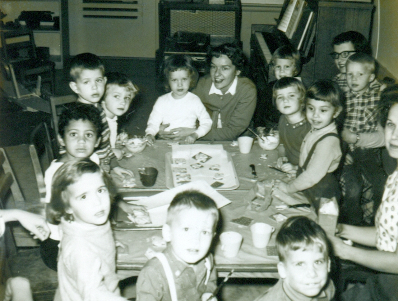 Old picture of children around a table looking at camera with teachers